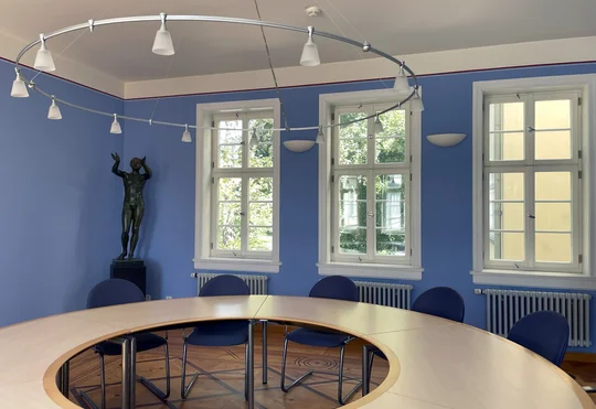 Ein Raum mit blauen Wänden, rundem Konferenztisch, einer Fensterfront. In der Ecke steht eine historische Bronzeskulptur, die einen betenden, nackten Jüngling im leichten Kontrapost darstellt.